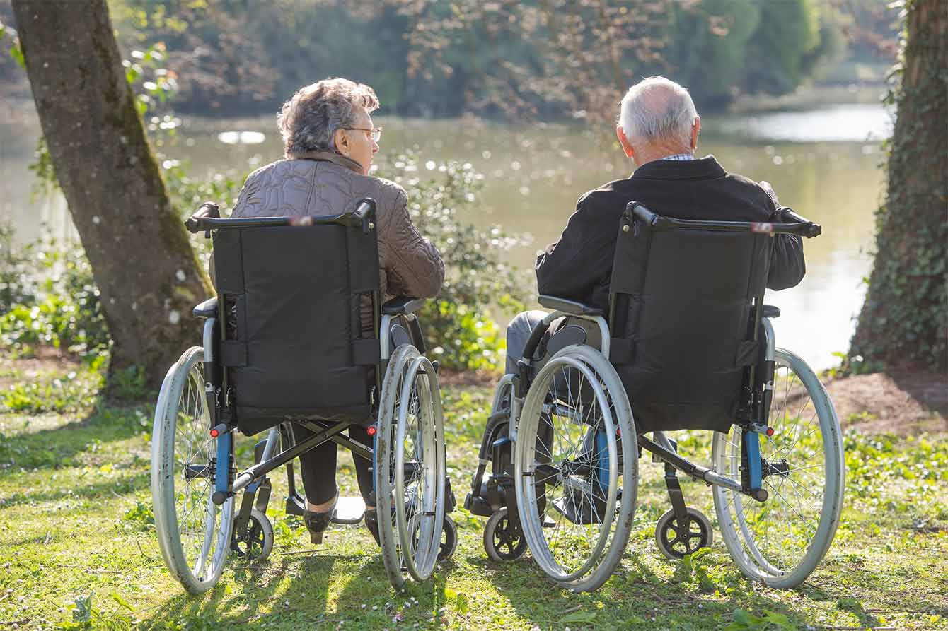 An couple in wheelchairs look at a lake, their backs towards us