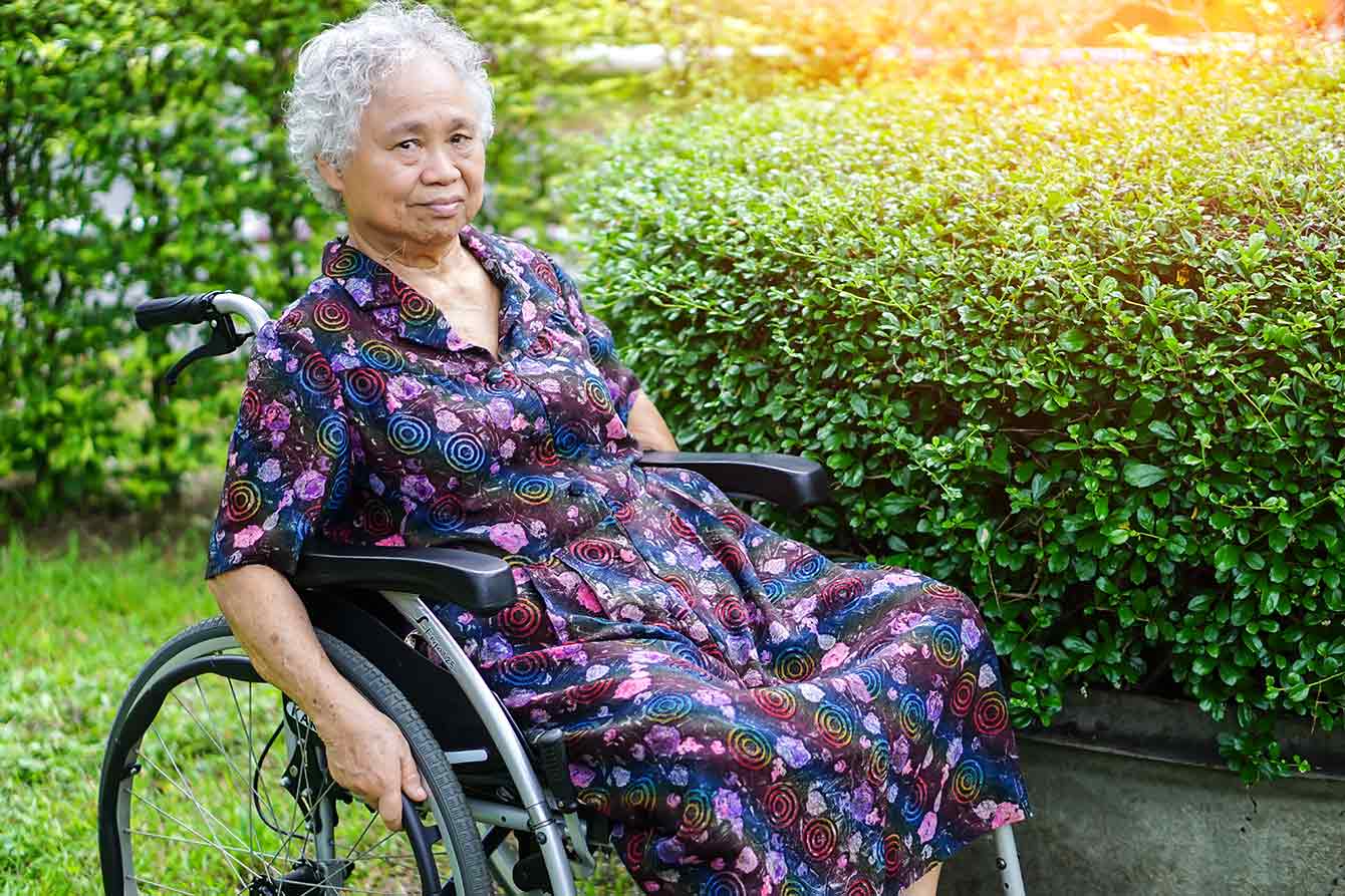 An elder Asian woman sits in wheelchair next to shrubbery