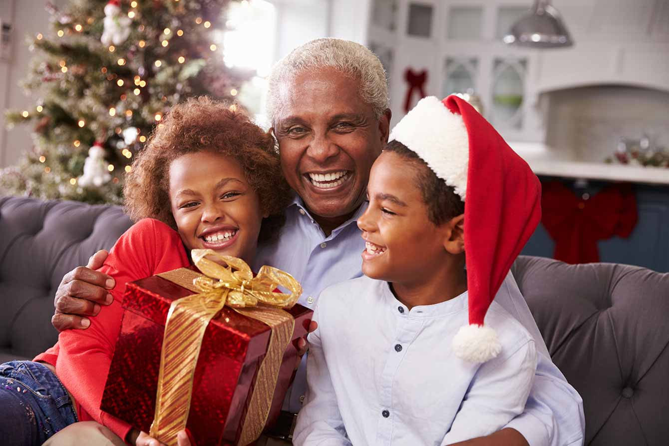 An older black man sits with grandchildren on couch with bow-wrapped present