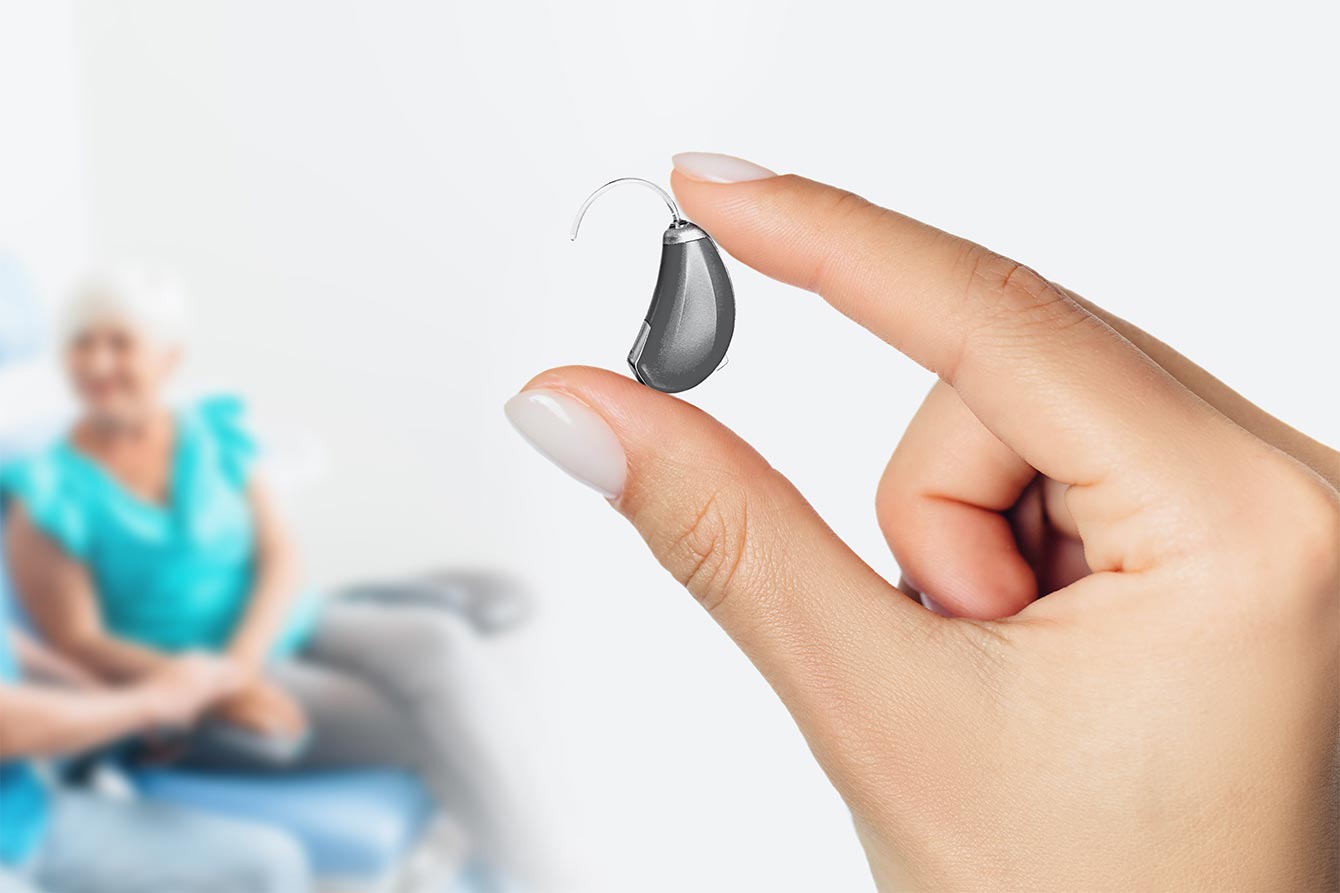 A hand holds a next gen hearing aid with an elderly couple waiting in the background