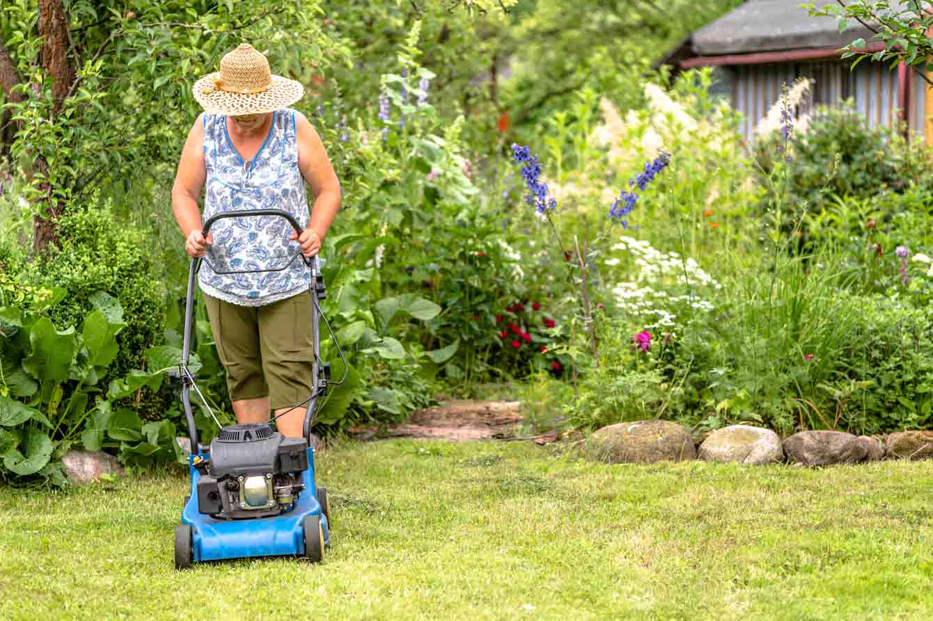 An older woman mows her lawn wearing a sun hat