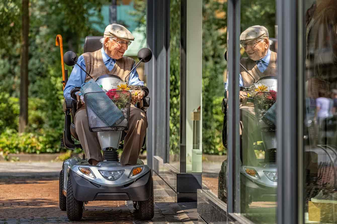 An elderly man rides a mobility scooter with a basket full of products while looking in a shop's window