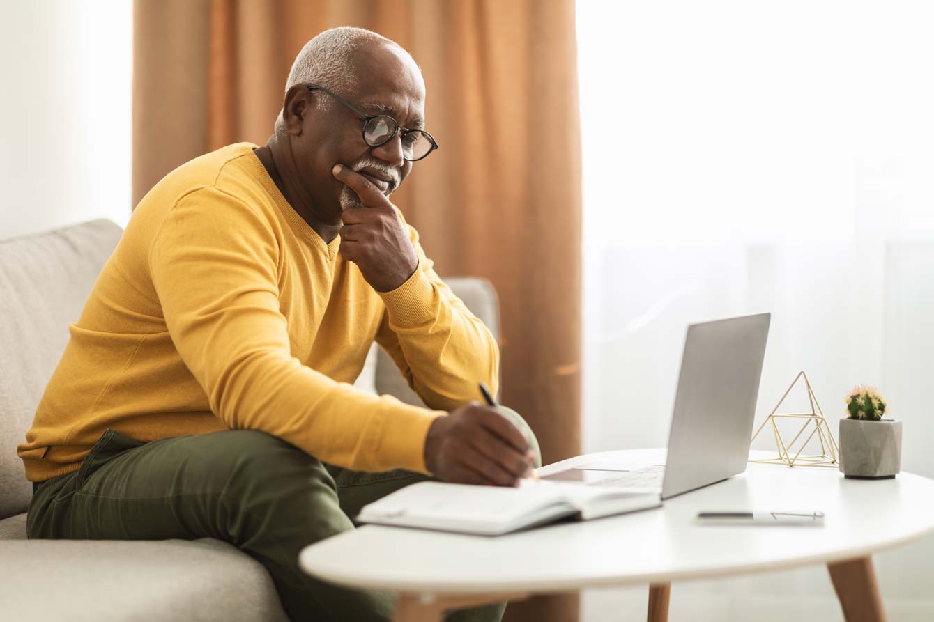 Older black man at laptop writing in a notebook taking an online class