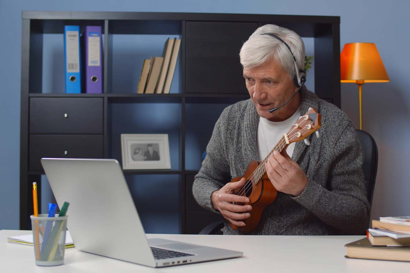 Senior man takes online music class while playing a ukulele in front of computer