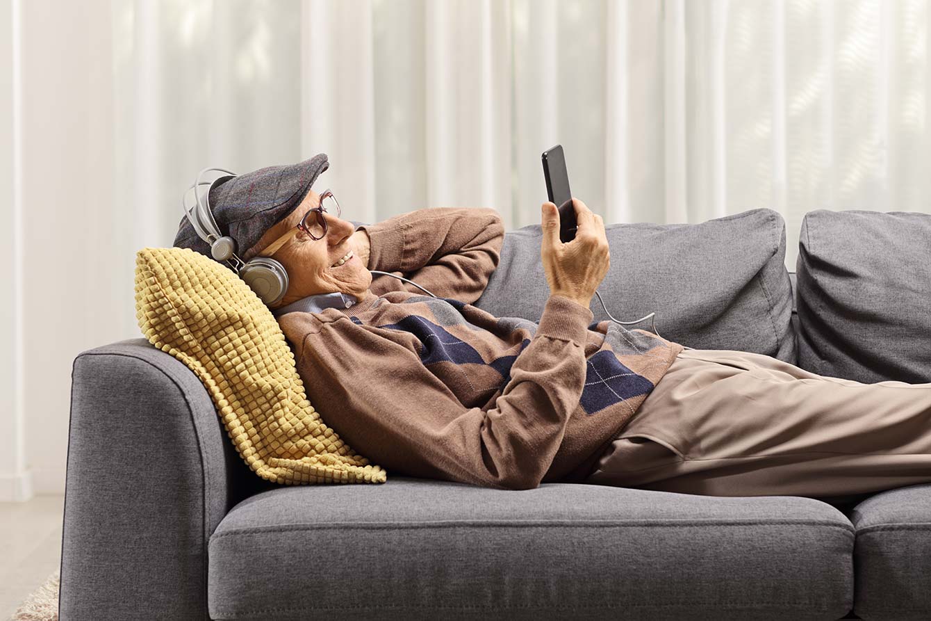 Elderly man on a sofa holding a mobile phone and listening to music on headphones