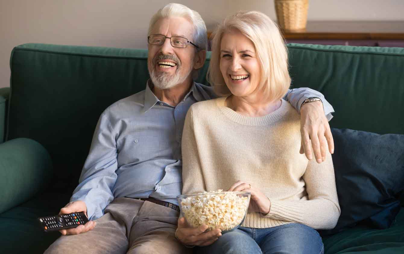 Happy older couple watch TV on couch while eating popcorn