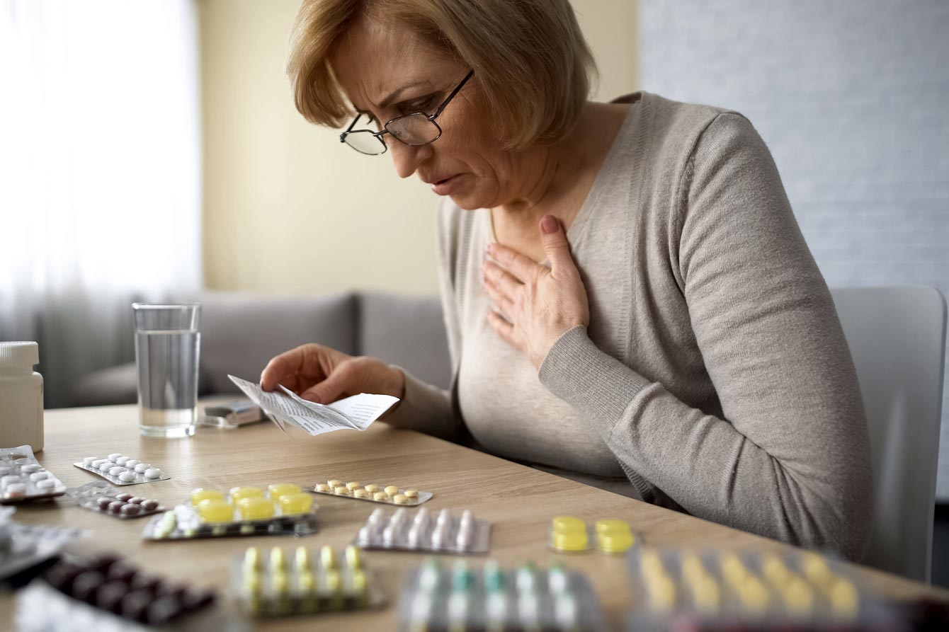 Older lady in front of a table covered with various medicines, reading instructions, unhappily