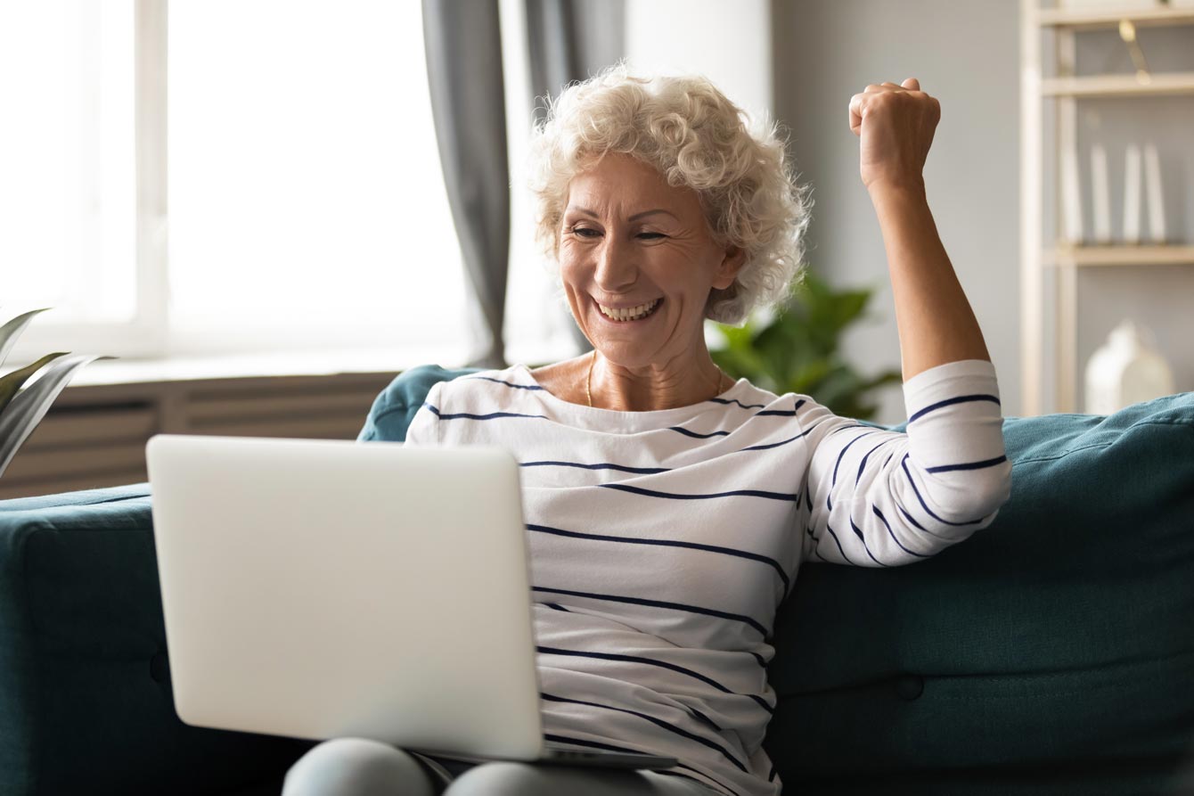 Old white woman sits on her couch with a laptop with her arm raised in the gesture of success