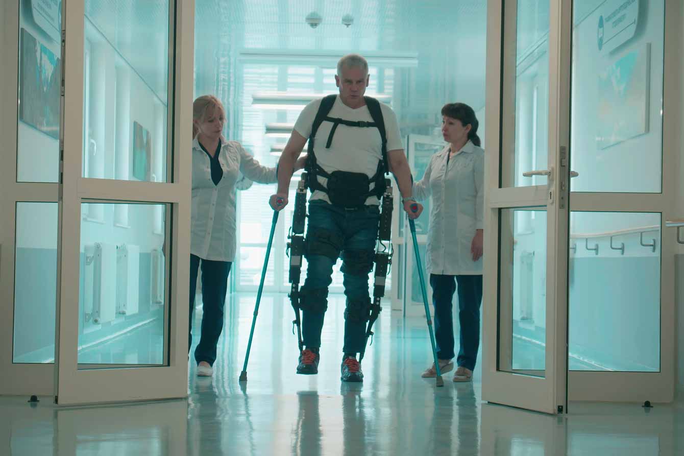 A senior in an exosuit walks with doctor support