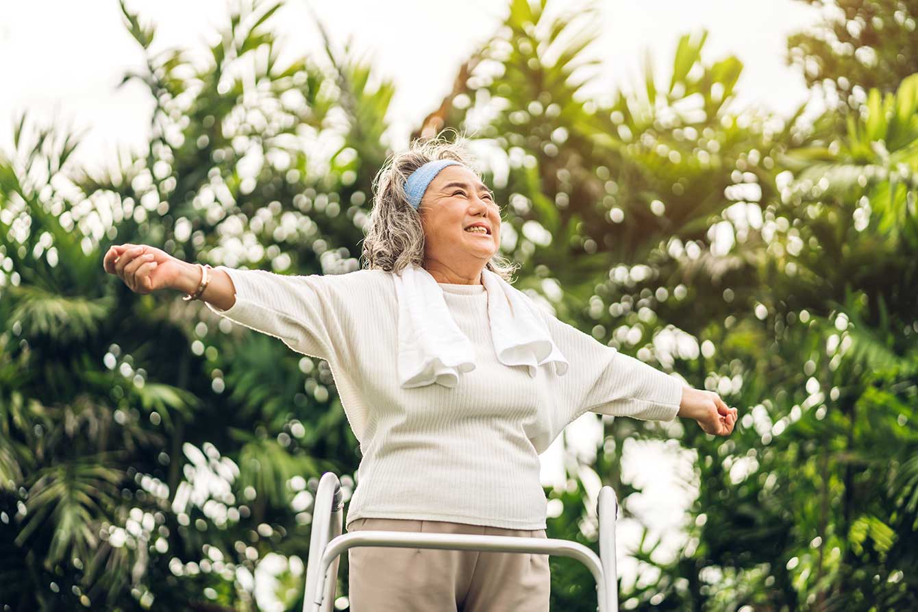 A happy Asian senior with walker stands with arms raised triumphantly in the park