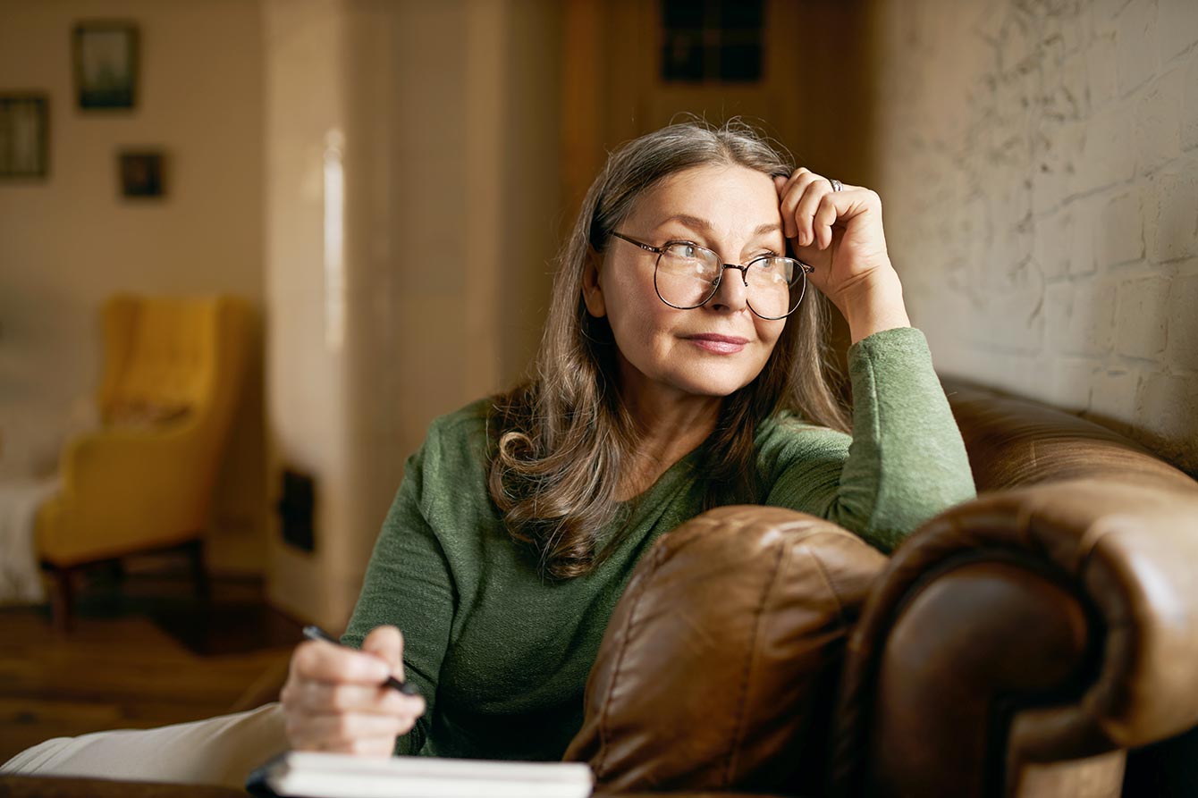 Older woman wearing glasses sits on couch journaling in a notebook