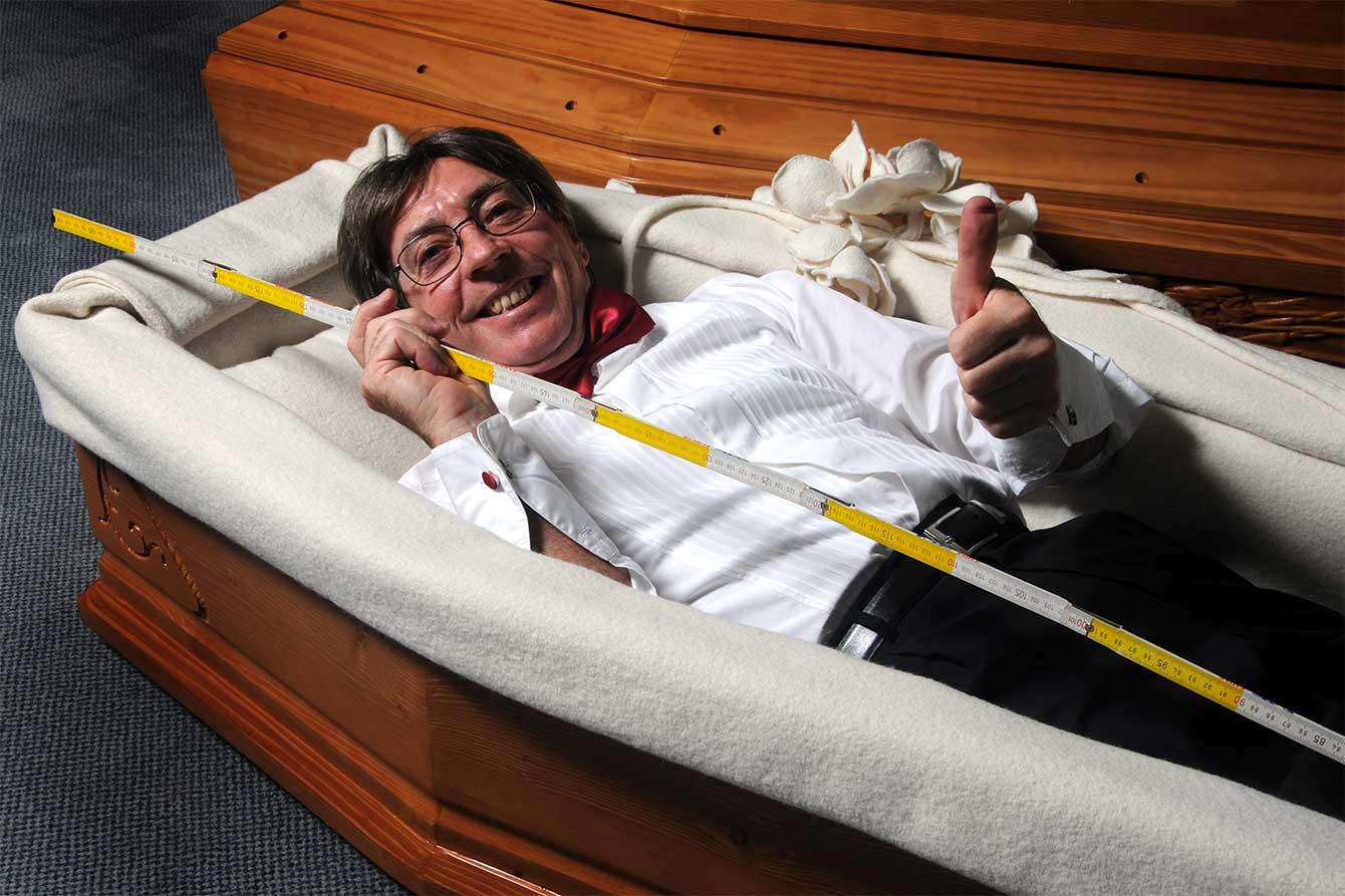 A smiling man measures himself with tape measure inside a funeral casket