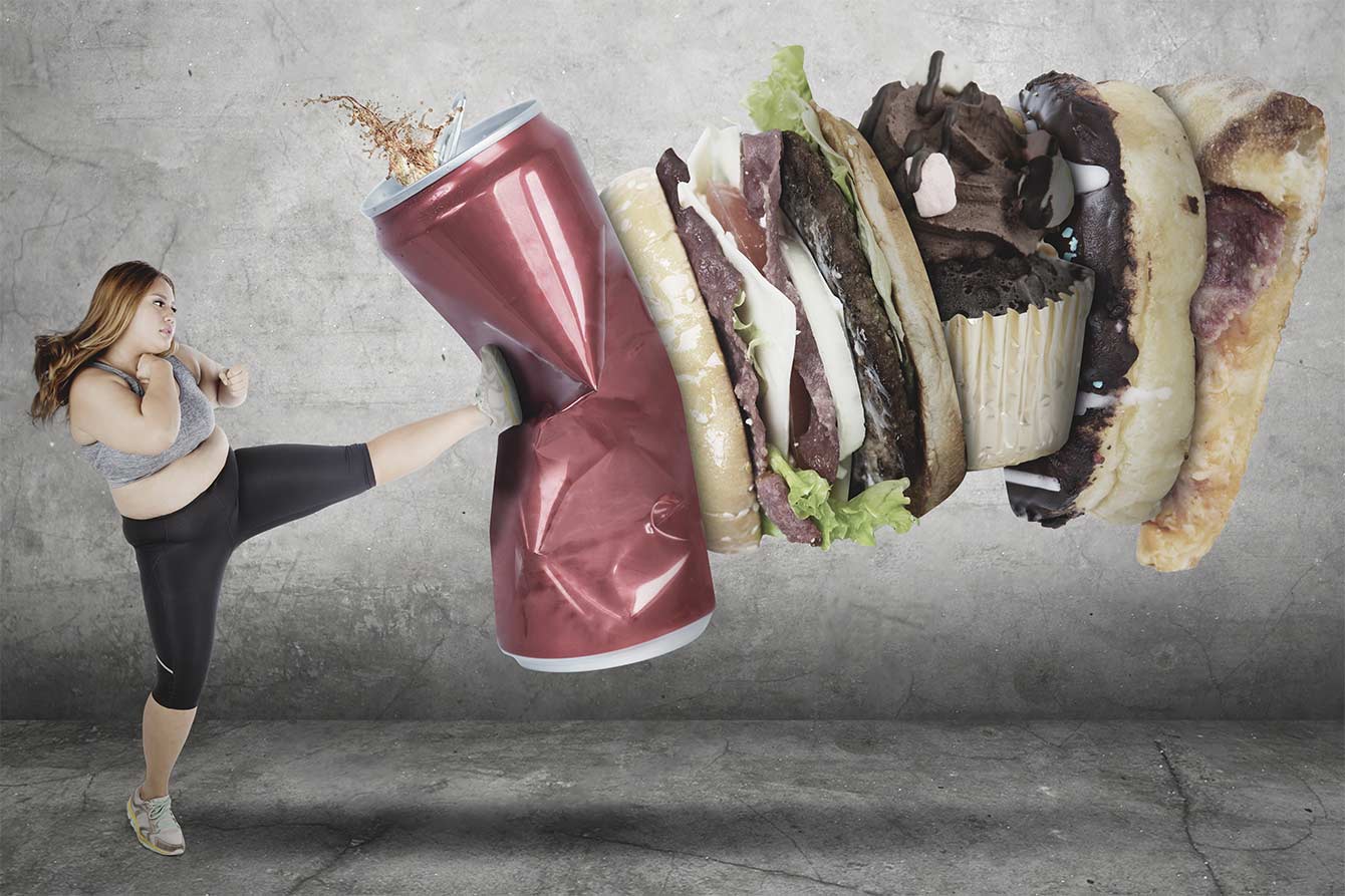 Overweight woman in exercise clothes jump-kicking unhealthy food and drink