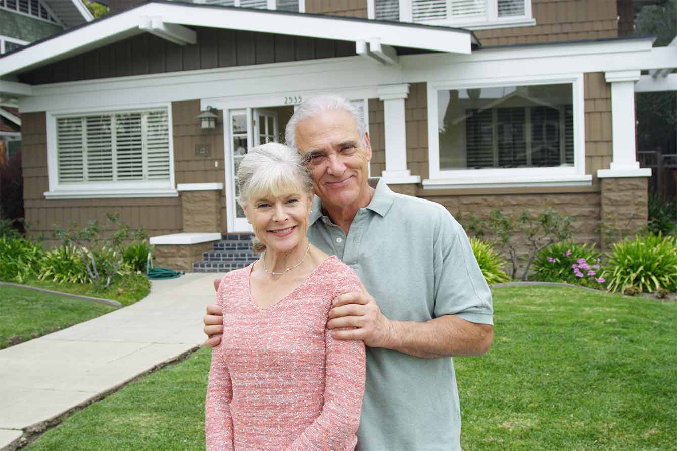 An older white couple stands in front of their small house