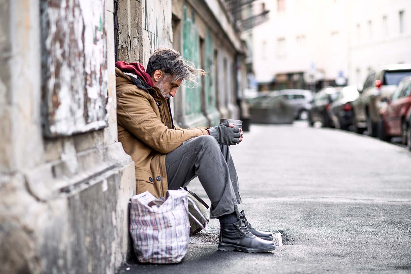 A homeless older man sits on the steps in front of a building in a city