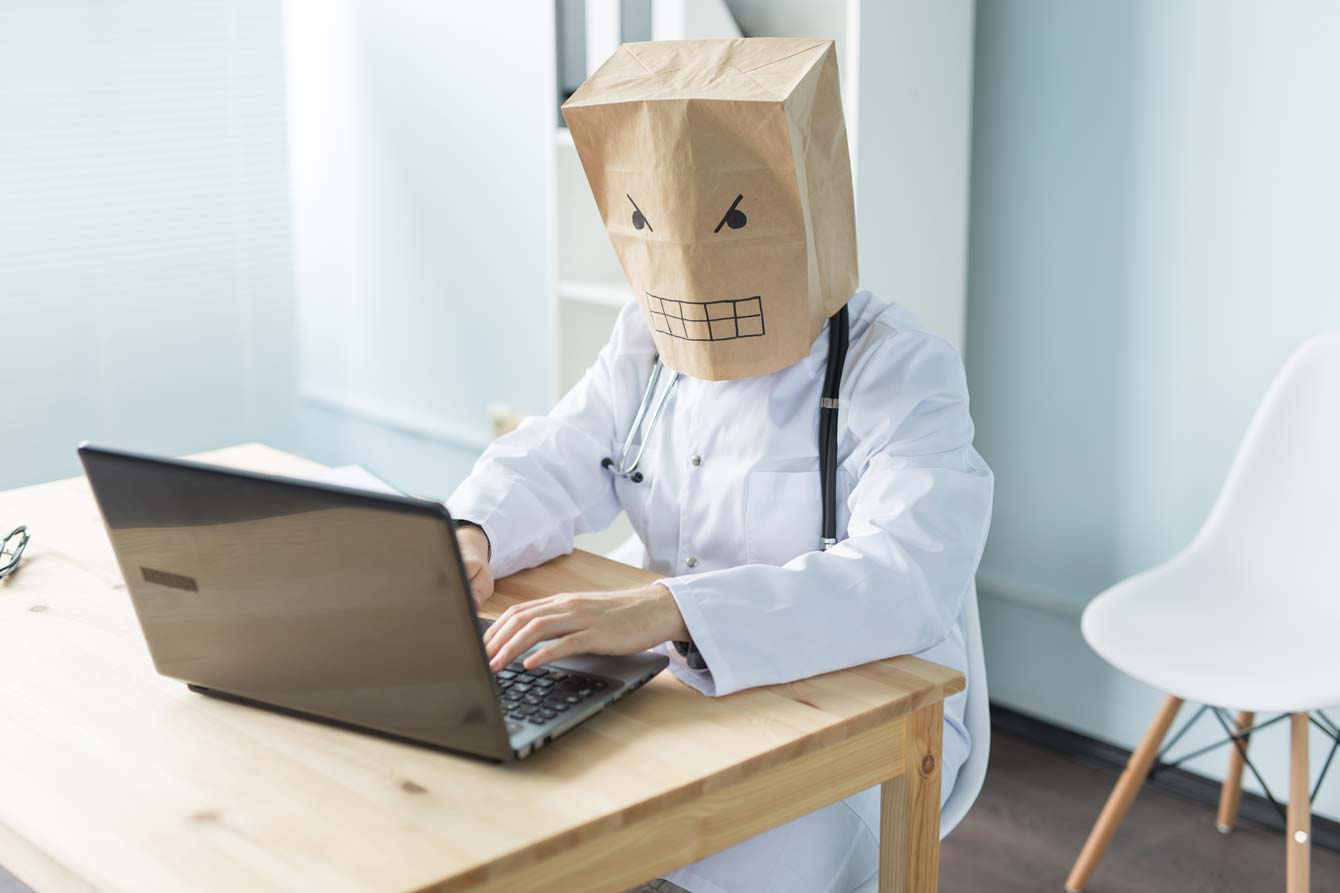 A doctor at a laptop with a paper bag over his had with an angry face drawn on to it