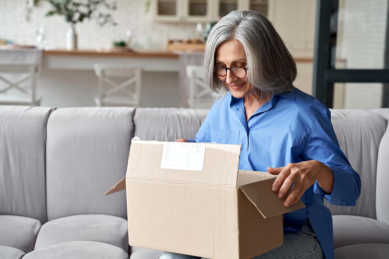 Older mature smiling woman sits on her couch opening a brown box
