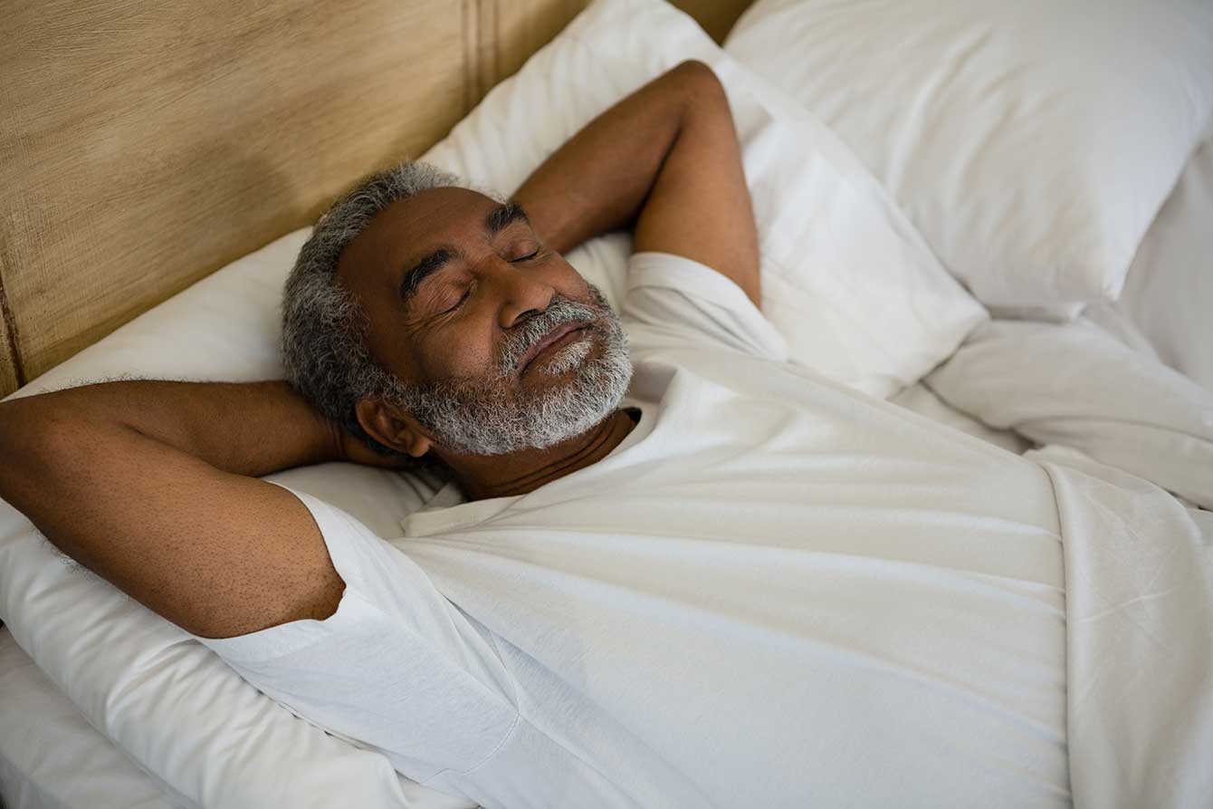 Elderly black man sleeping comfortably in bed with arms behind head
