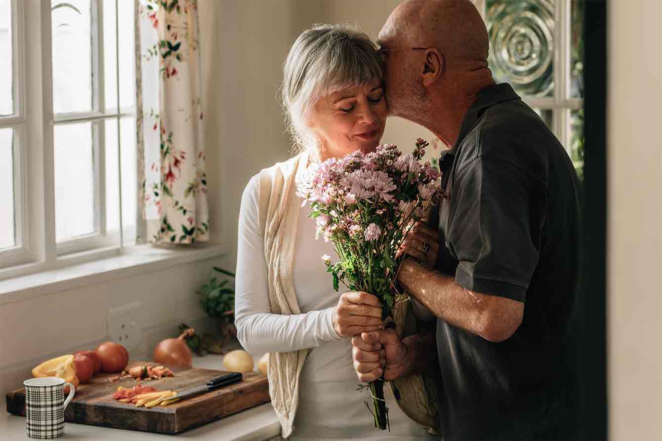 Senior romantic couple at home in kitchen giving flowers