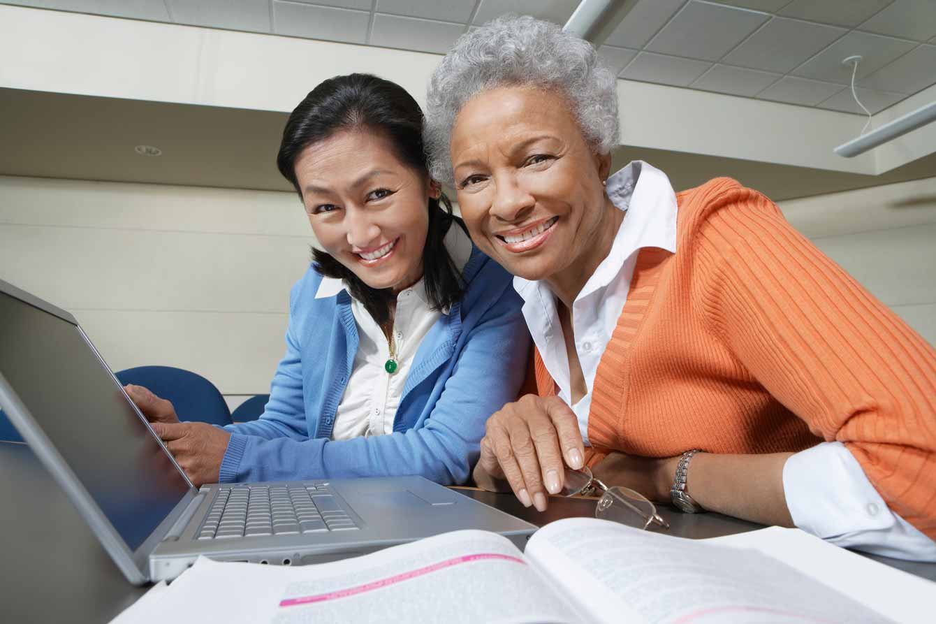 An elderly black woman sits at a laptop next to an Asian woman