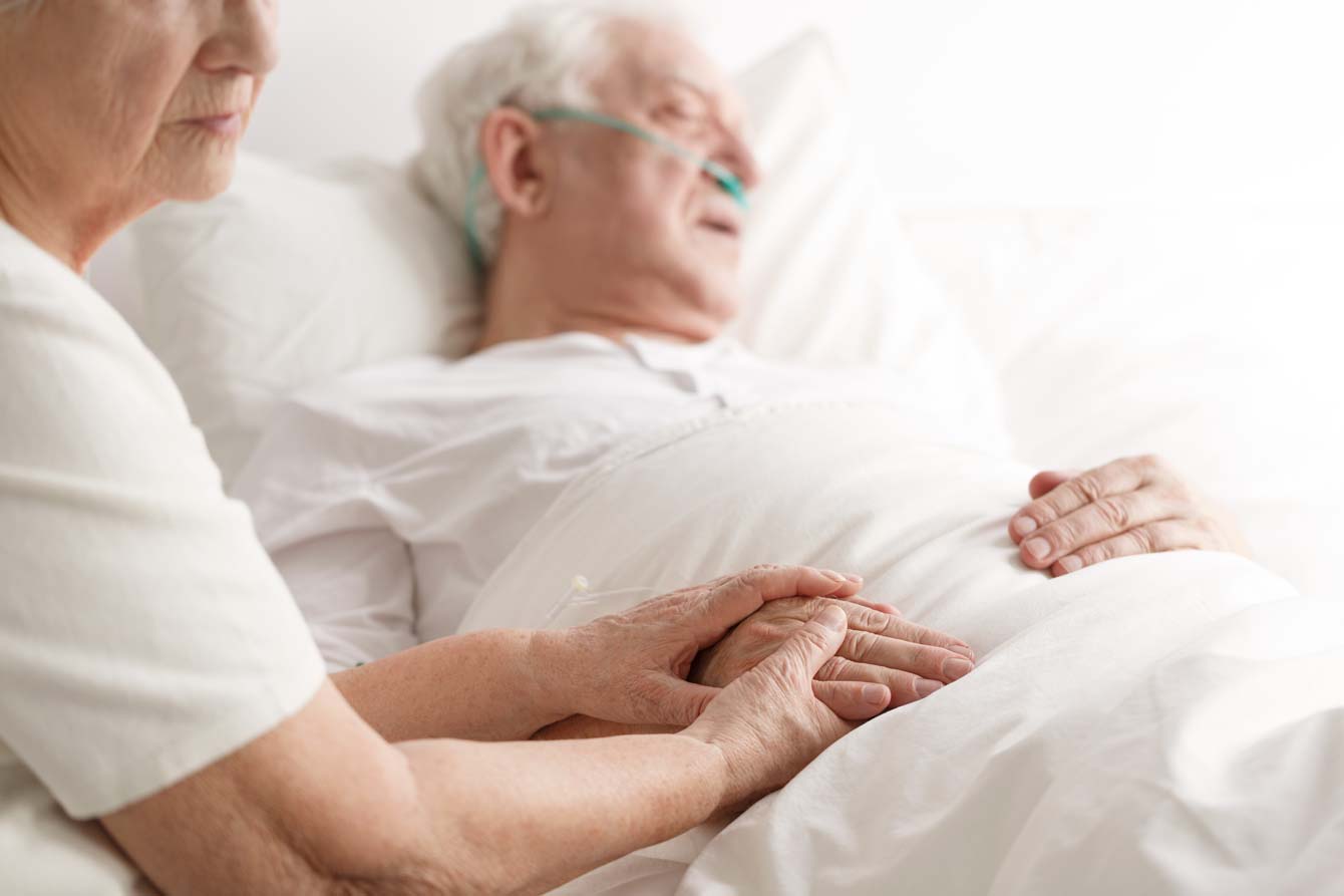 An older woman holds the hand of her dying husband who sleeps in hospital bed with oxygen tubes in nose