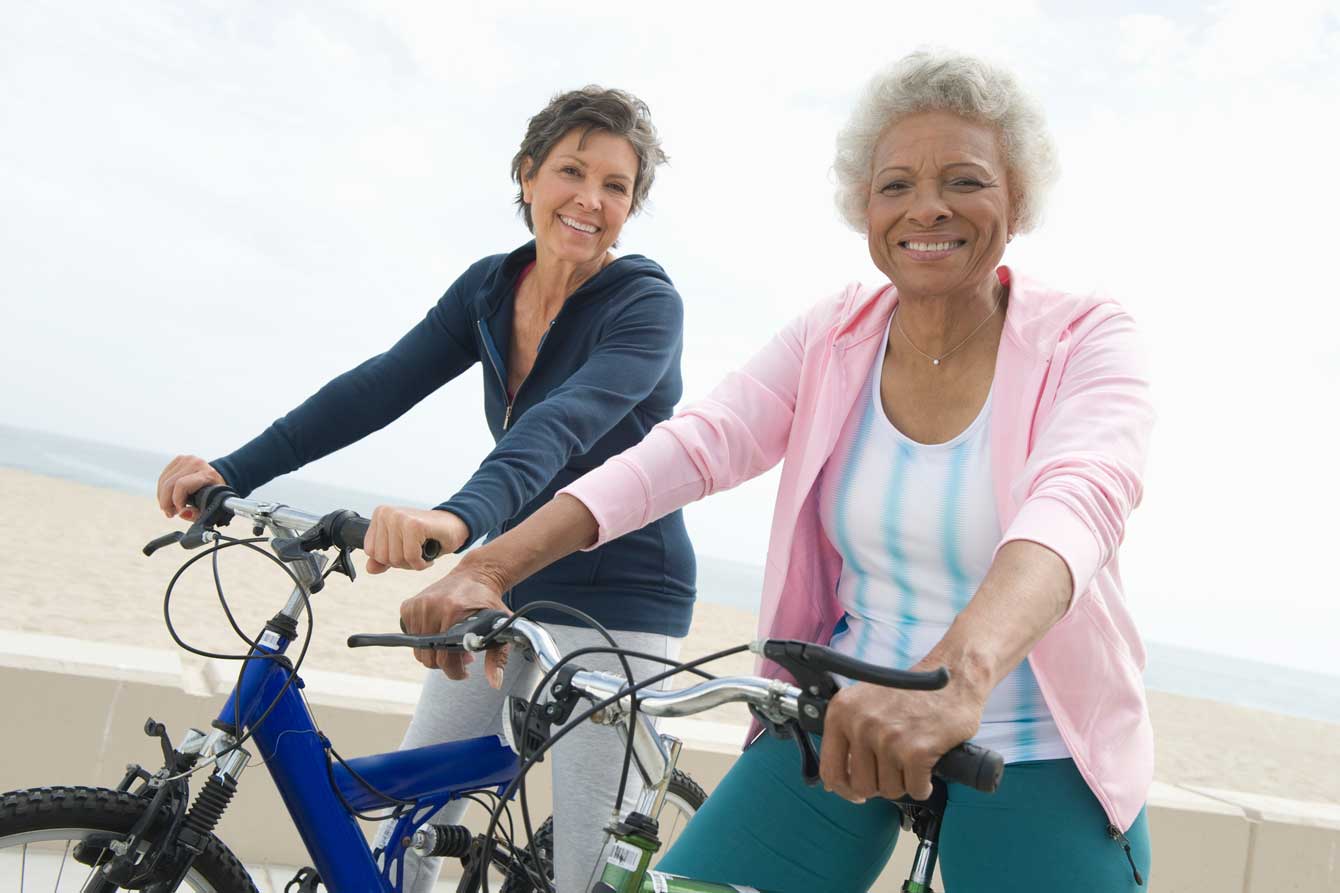 Two older ladies ride bikes together on the beach