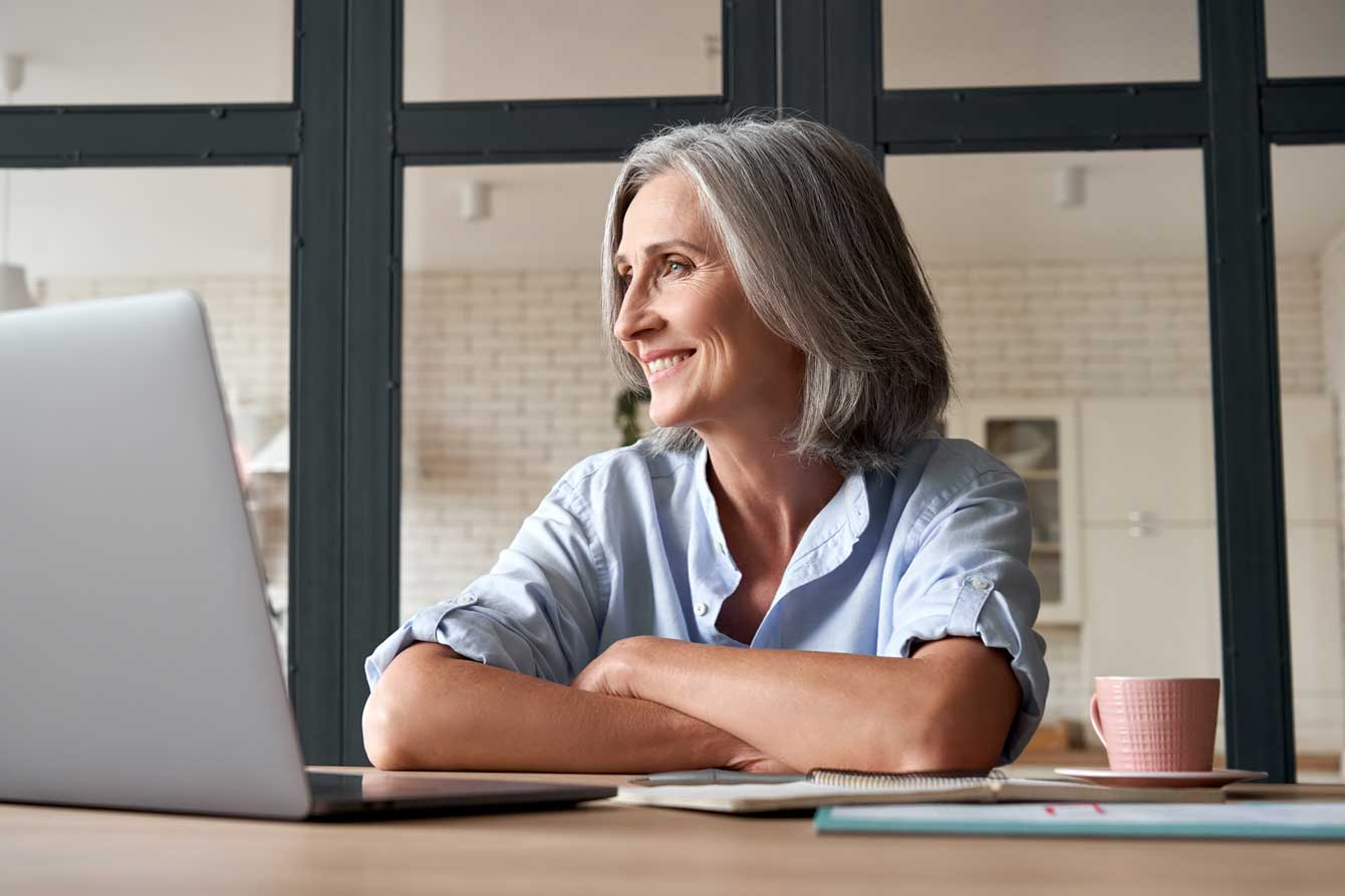 An attractive smiling older woman with gray hair sits in front of her laptop