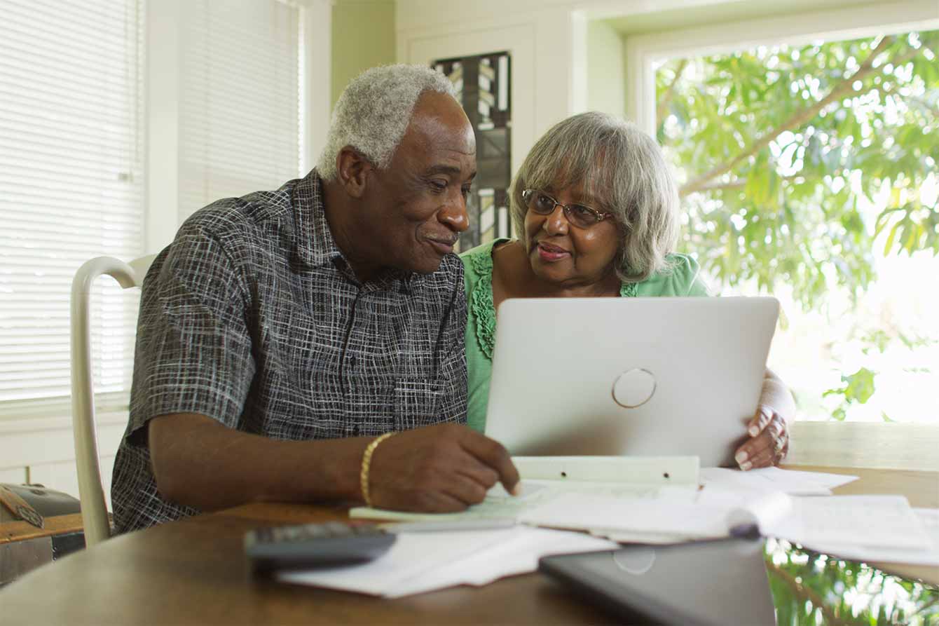 An elderly black couple review bills on a table in front of a laptop computer
