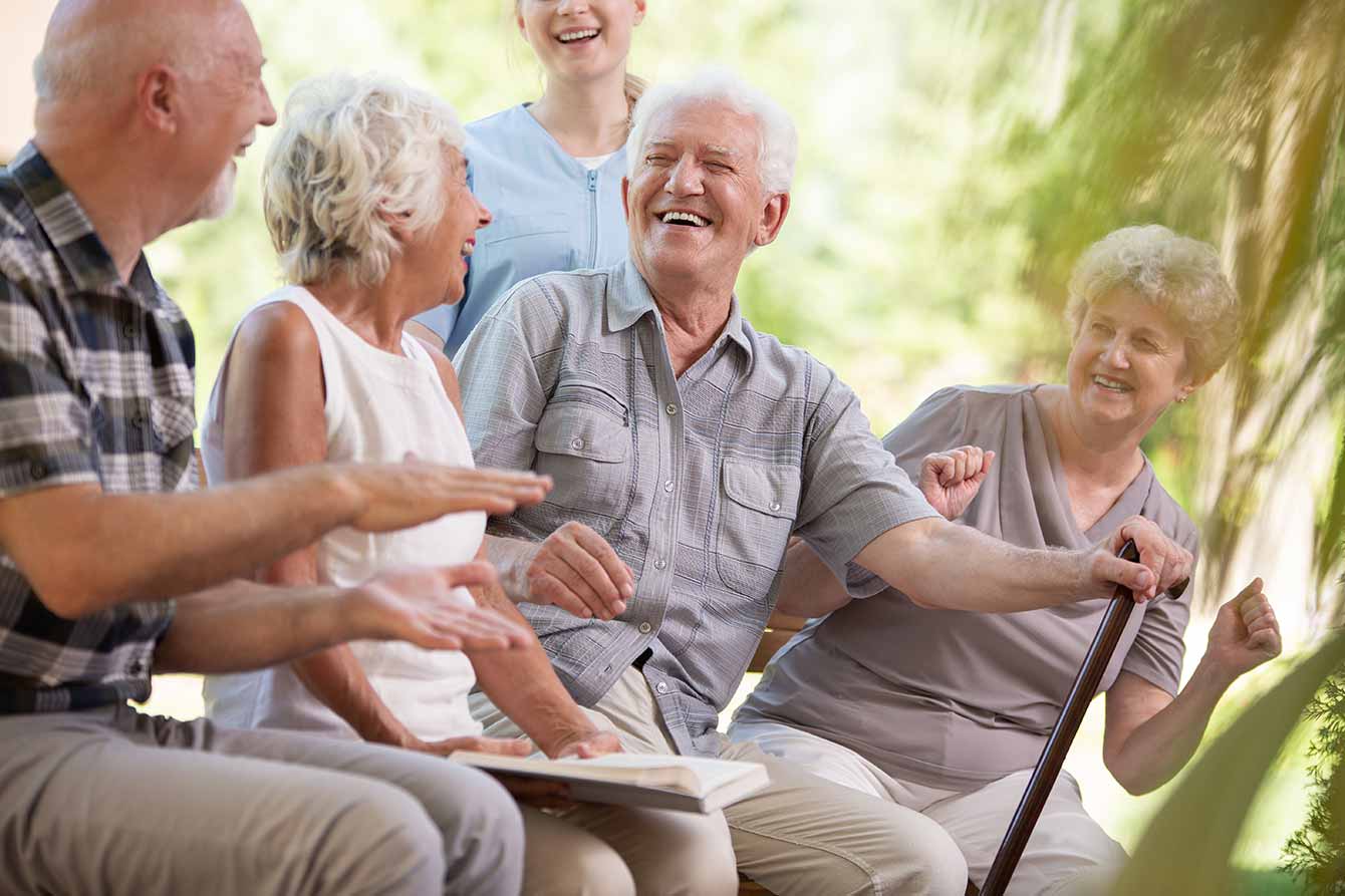A group of senior friends laugh together while sitting on a bench in a park
