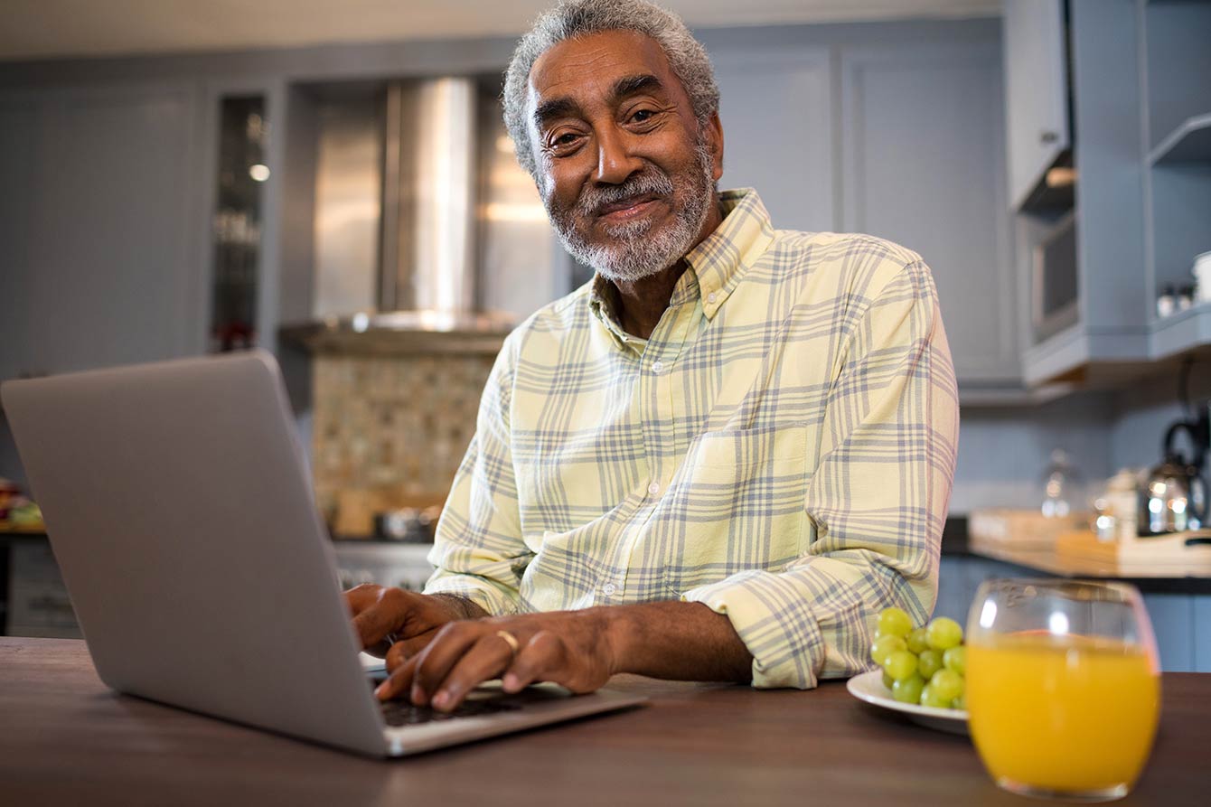 Male senior sitting at desk using laptop with orange juice and grapes next to him