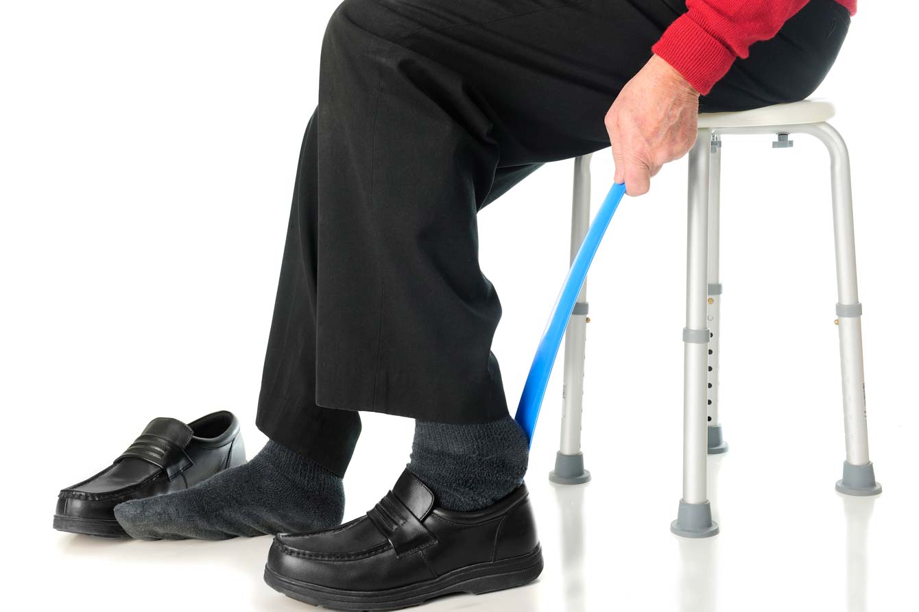 An older man uses a shoe horn to put on his shoes while sitting on a stool