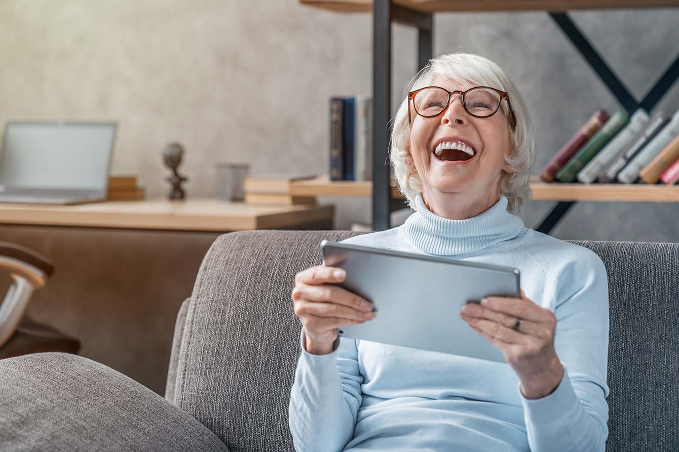 Happy senior woman looking and laughing at her digital tablet on sofa