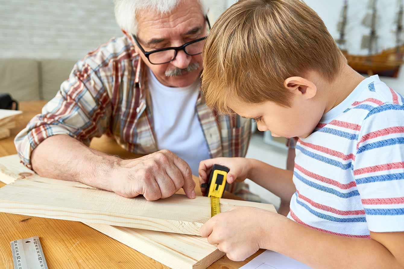 Senior man helping little boy build birdhouse teaching him woodwork at table in studio