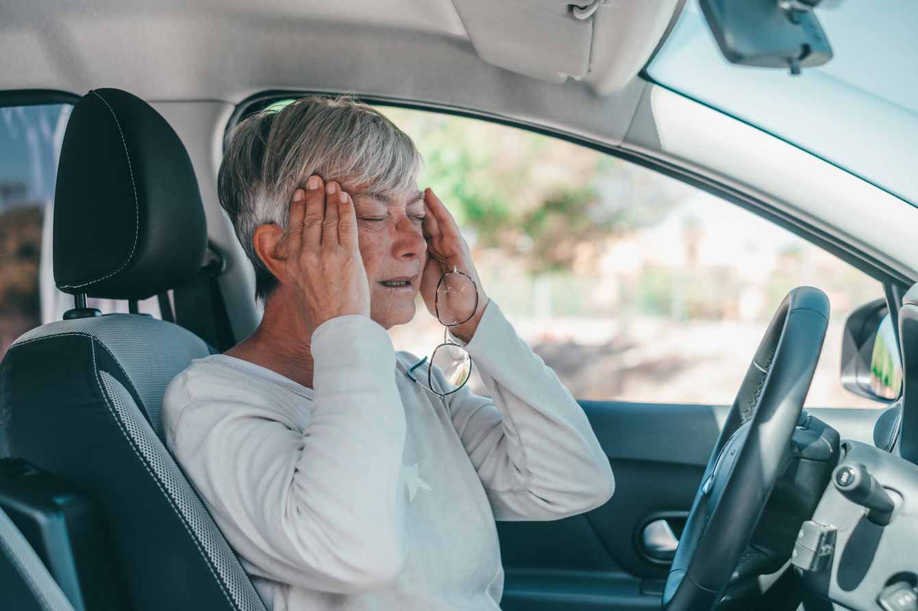 A senior female in her car rubs her temples with a painful expression on her face