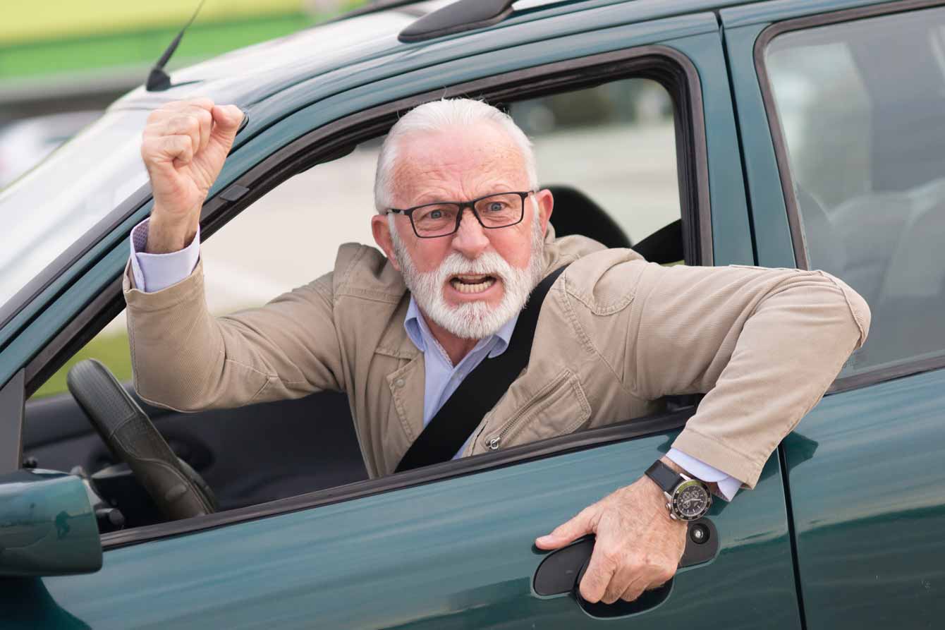 An angry senior man in his car angrily shakes his fist out the car window at another driver
