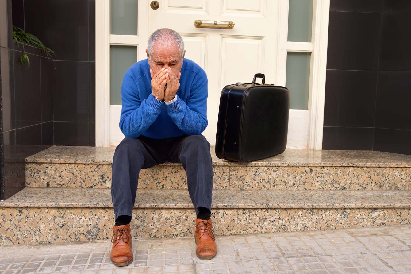senior man downtrodden by the porch of his home with his suitcase